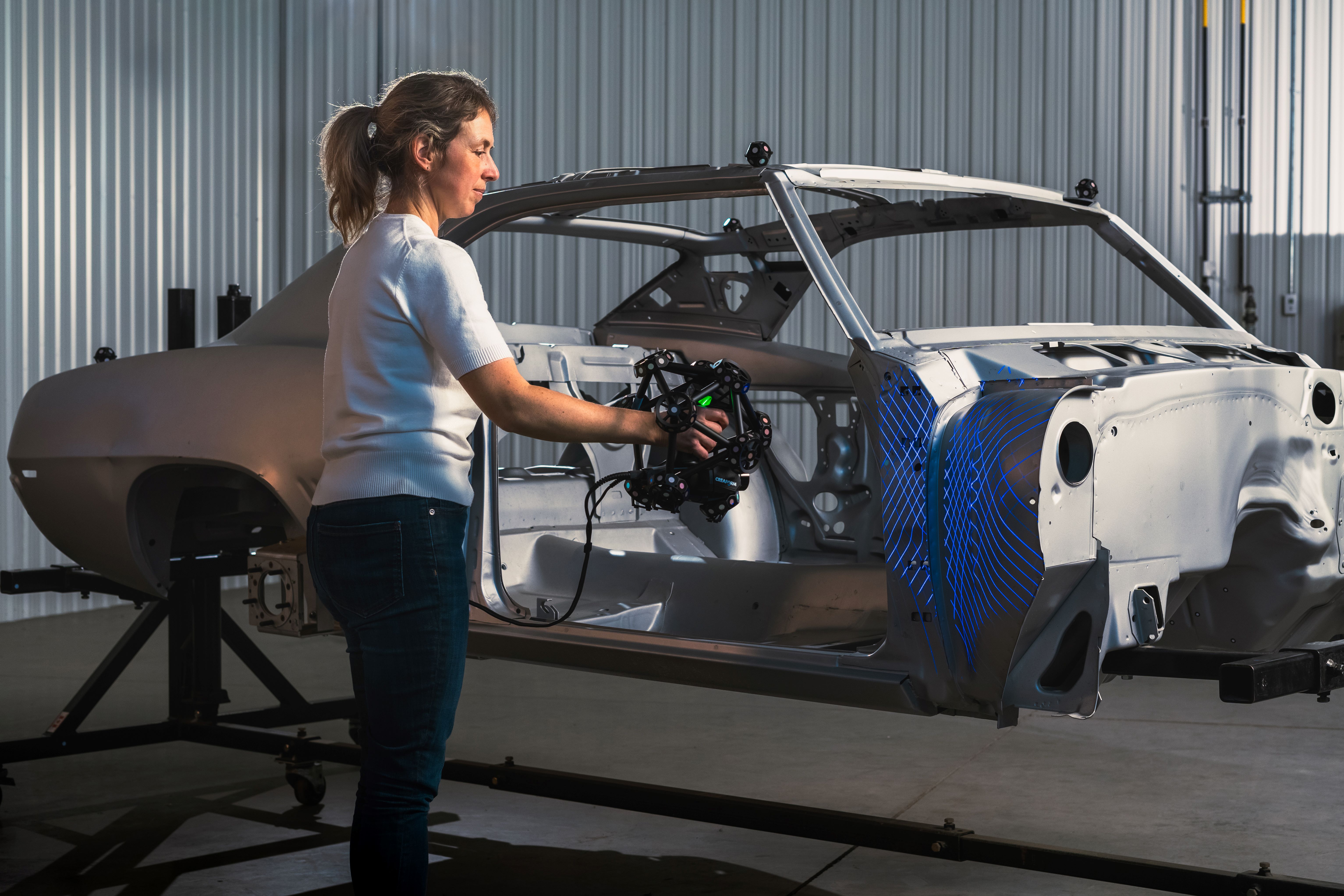 A woman in a warehouse using the MetraSCAN 3D to measure a grey car frame mounted on a support frame.