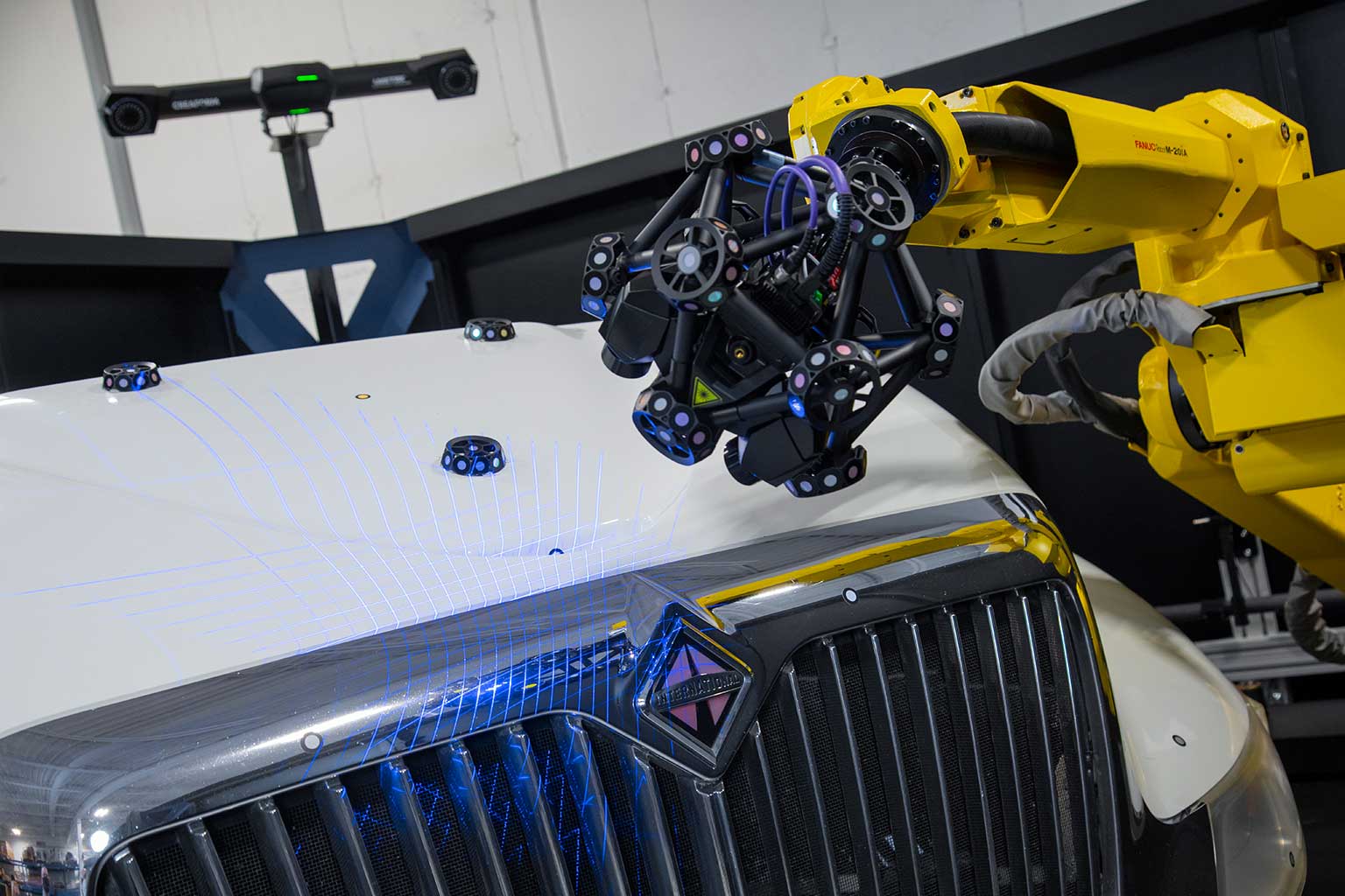 CMM óptica de MetraSCAN 3D montado en robot de la R-Series escaneando un parachoques y un capó de camión internacional blanco con láser azul y C-track en el fondo